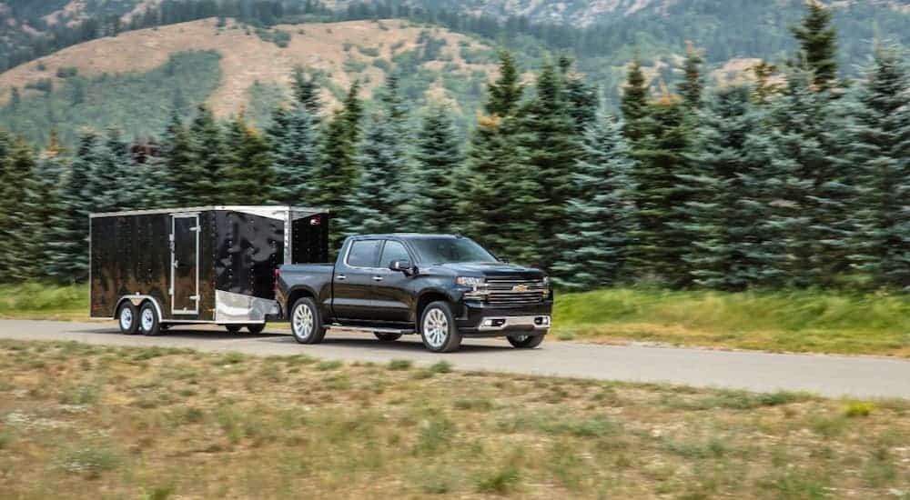 A black 2021 Chevy Silverado 1500 High Country is towing a black enclosed trailer in front of evergreen trees.