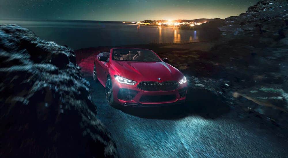 A red 2021 BMW M8 convertible is shown from the front driving on a coastal road at night.