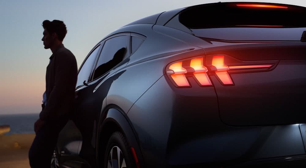 The silhouette of a man is shown leaning on a gray 2021 Ford Mustang Mach-E with its taillights illuminated.