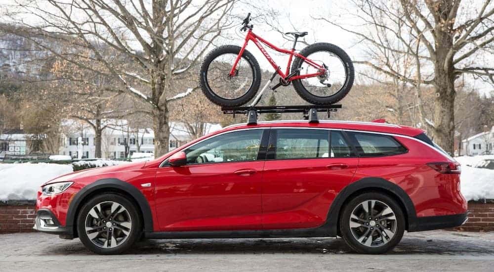 A red 2019 Buick Regal TourX is shown from the side with a bike on the roof parked with a snowy town in the background.