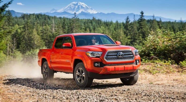 A red 2016 Toyota Tacoma is driving on a dirt trail in front of trees and mountains.