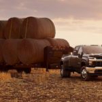 A black 2021 Chevy Silverado 3500 HD is parked in a field with a trailer of hay bales at sunset.