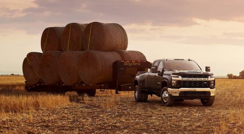 A black 2021 Chevy Silverado 3500 HD is parked in a field with a trailer of hay bales at sunset.