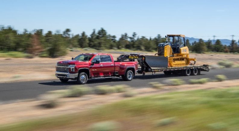 A red 2021 Chevy Silverado 3500 HD is towing heavy machinery on a rural road.