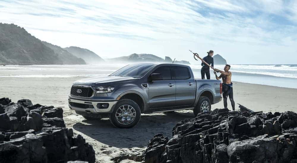 A grey 2021 Ford Ranger is parked on the beach as two people prepare to surf. 
