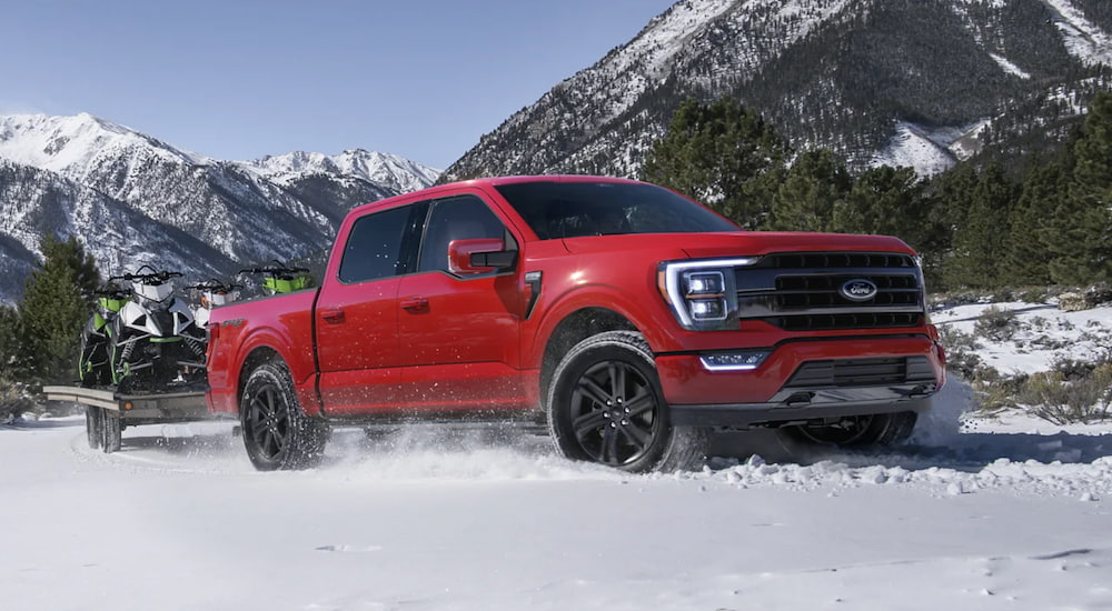 A red 2021 Ford F-150 is driving through the snow while pulling snowmobiles.