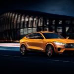An orange 2021 Ford Mustang Mach-E is driving through a city at night after leaving a Ford Mustang Mach-E dealer.