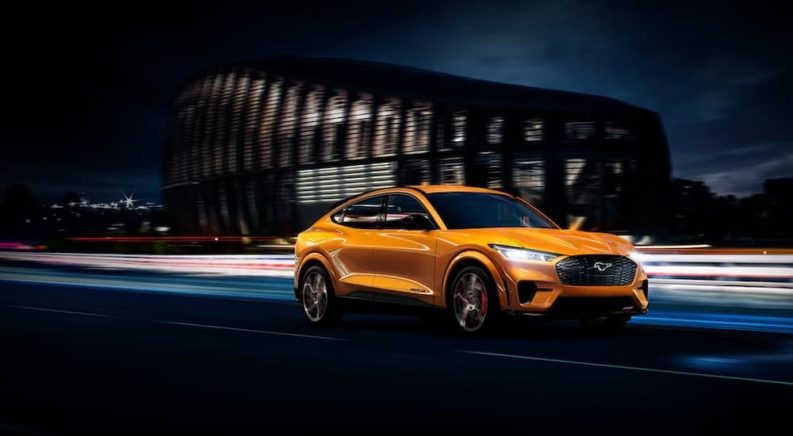 An orange 2021 Ford Mustang Mach-E is driving through a city at night after leaving a Ford Mustang Mach-E dealer.