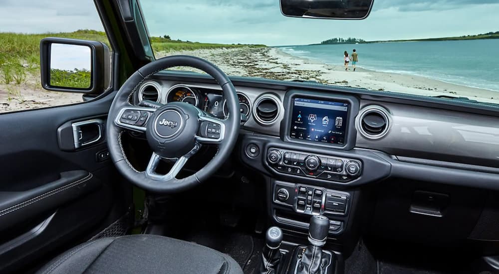 The steering wheel and infotainment screen of a 2021 Jeep Wrangler is shown parked on a beach.