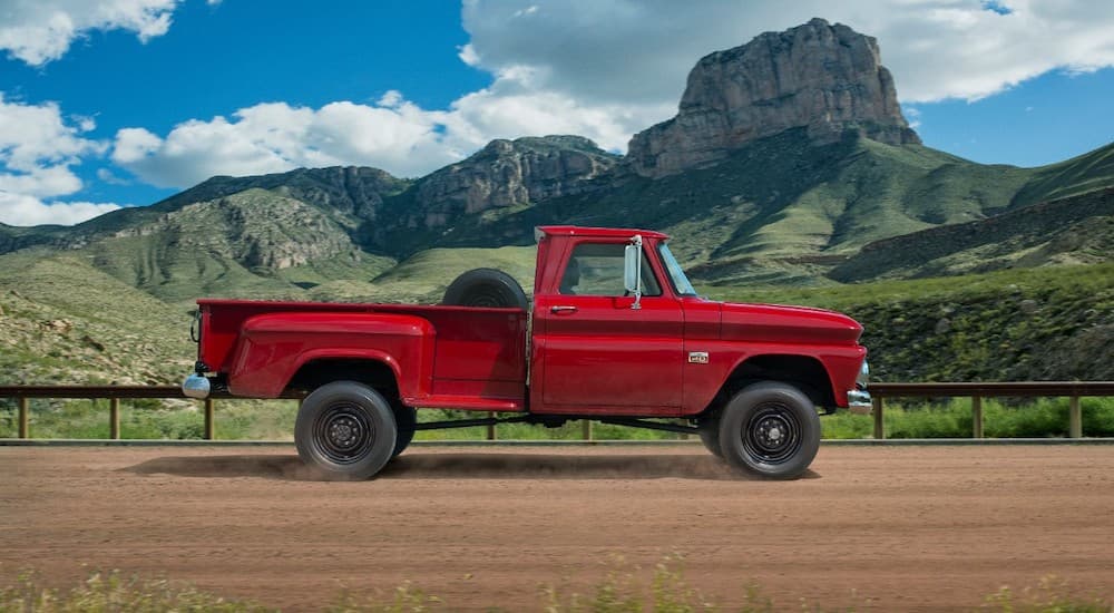 A red 1960s Chevy C/K series truck is driving on a dirt road past mountains.