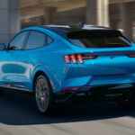 A light blue 2021 Ford Mustang Mach-E is driving on a road after leaving a local Ford SUV dealer.