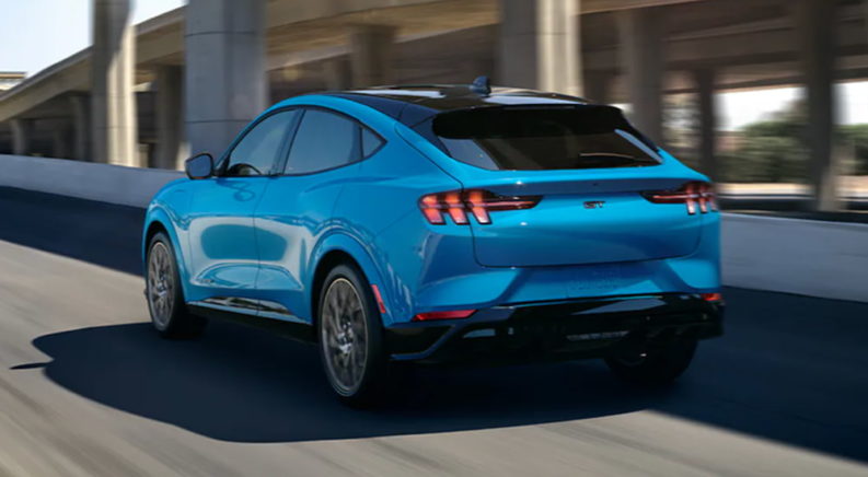 A light blue 2021 Ford Mustang Mach-E is driving on a road after leaving a local Ford SUV dealer.
