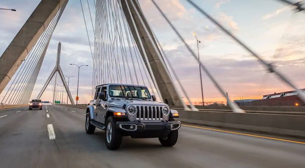 A silver 2021 Jeep Wrangler is driving over a bridge at sunset. 