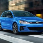 A blue 2021 Volkswagen Golf GTI is show from the front driving through a city.