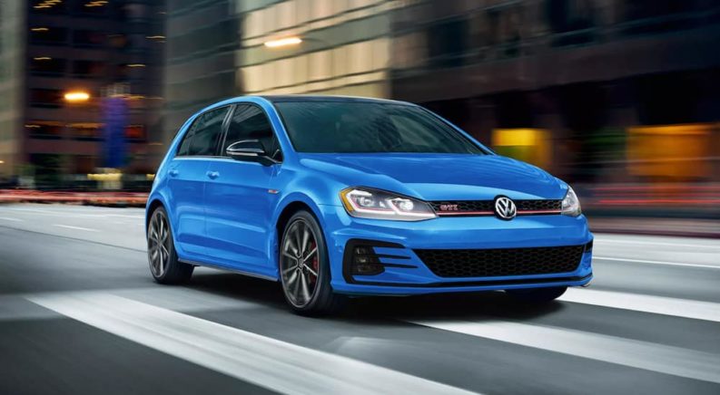 A blue 2021 Volkswagen Golf GTI is show from the front driving through a city.