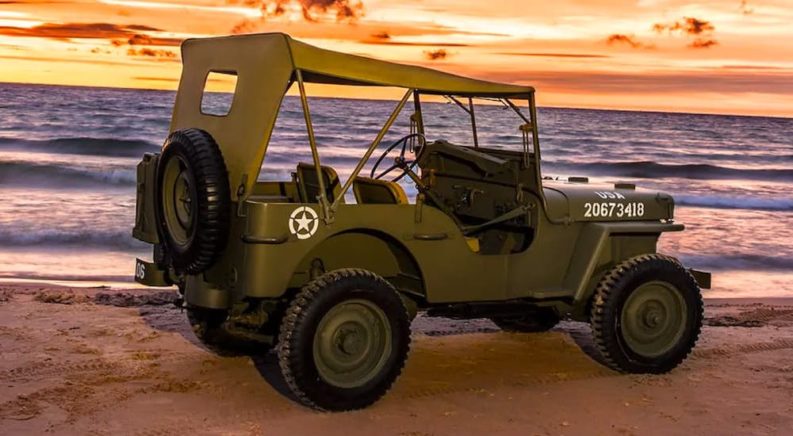 A green 1942 Jeep Wrangler Willys MB is shown from the side on a beach at sunset.