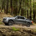 A grey 2022 Ford F-150 Lightning Lariat is shown from the side driving on a dirt path in the woods.