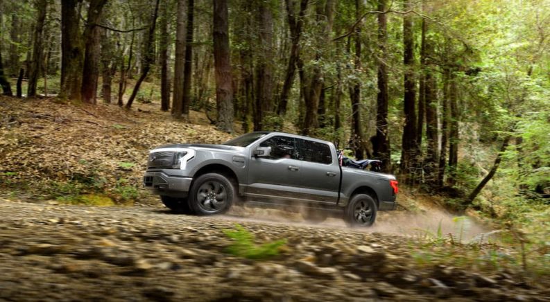 A grey 2022 Ford F-150 Lightning Lariat is shown from the side driving on a dirt path in the woods.