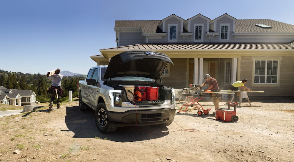 A silver 2022 Ford F-150 Lightning Pro is shown with the frunk open while a building a house.
