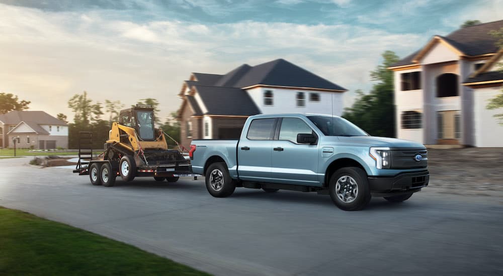 A pale blue 2022 Ford F-150 Lightning Pro is shown towing construction equipment past suburban houses.