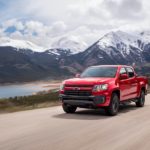 A red 2022 Chevy Colorado Trailboss is shown driving on a dirt road past a lake and mountains.