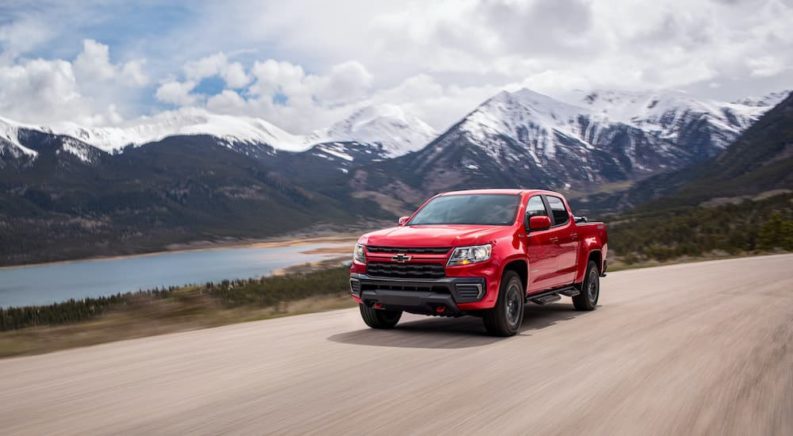 A red 2022 Chevy Colorado Trailboss is shown driving on a dirt road past a lake and mountains.