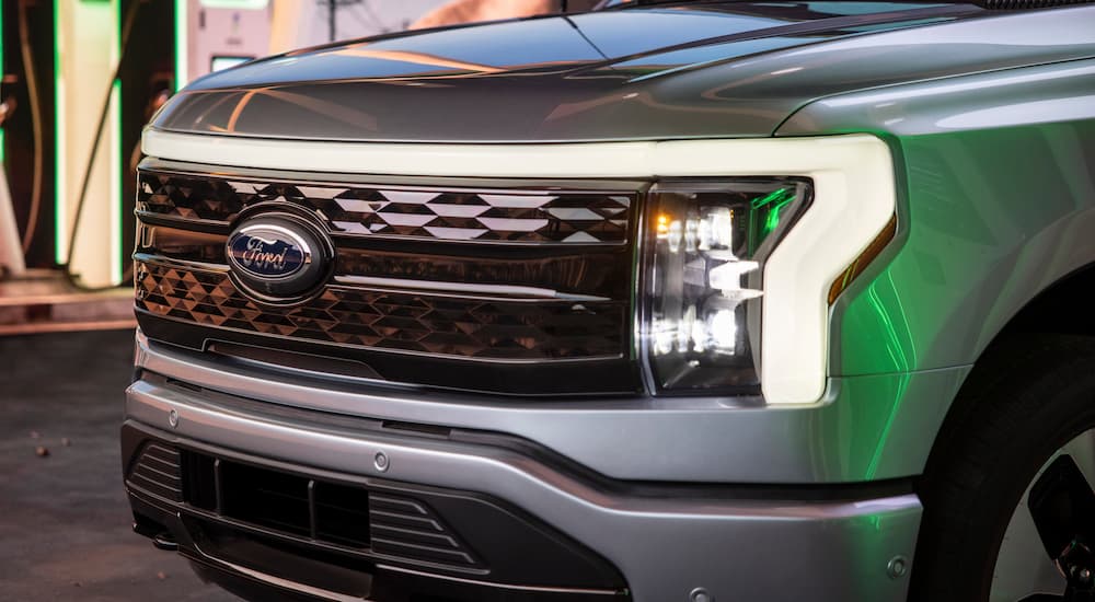 A close up of the grille of a silver 2022 Ford F-150 Lightning is shown with the headlights on.