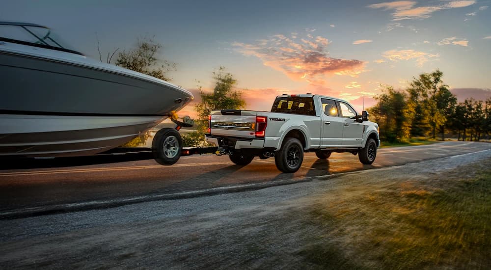 A white Ford F-250 Platinum is shown towing a boat on a road near a lake.
