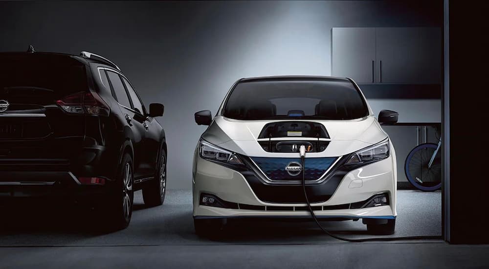 A white 2022 Nissan LEAF is shown charging in a garage next to a black Nissan Rogue.
