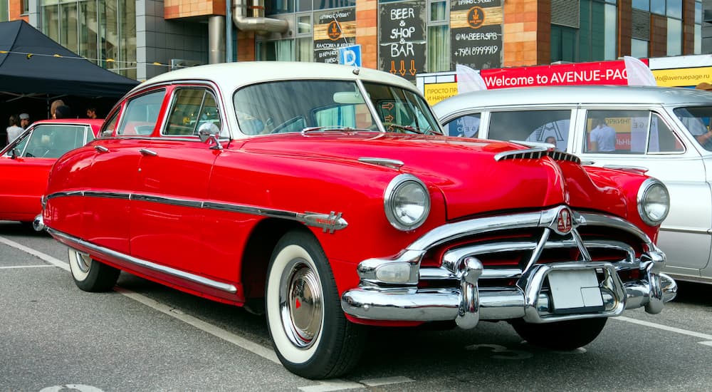 A red 1950s Hudson Hornet is shown parked in a city lot next to a silver vehicle.