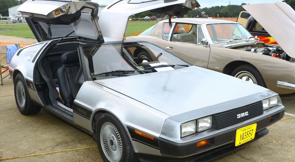 A silver 1980s Delorean is shown parked in grass with it's driver and passenger doors open.
