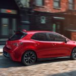 A red 2021 Toyota Corolla Hatchback is shown from the side driving on a city street.