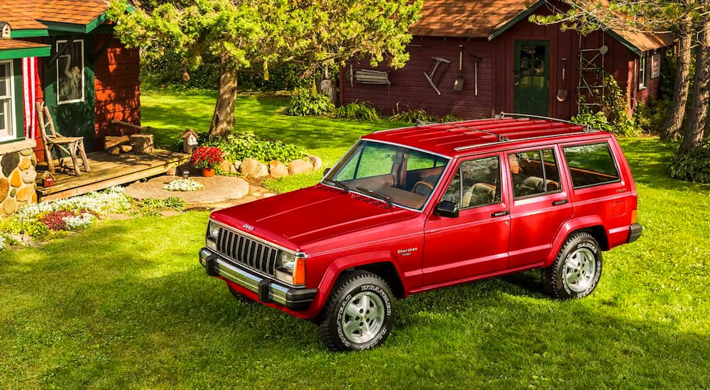 A red 1987 Jeep Cherokee XJ is shown parked in the grass in front of a cabin.