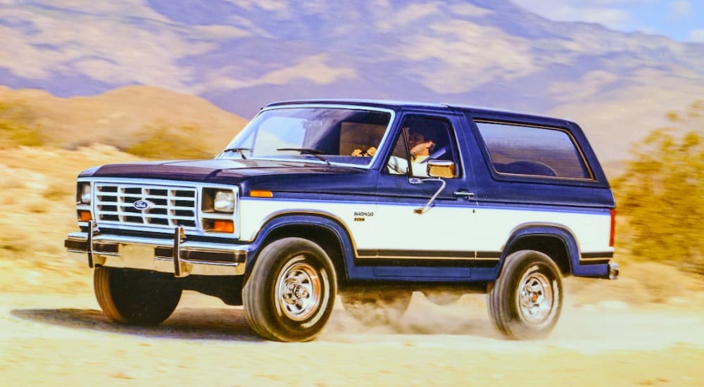 A blue and white 1986 Ford Bronco is shown driving on a dusty road.