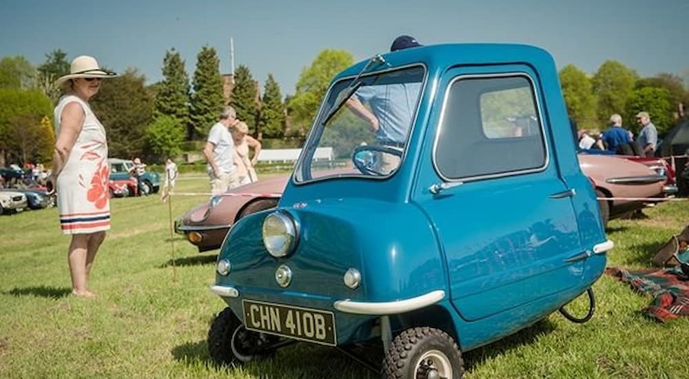 A blue 1962 Peel P50 is shown parked in a grassy field at a car show