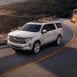 A silver 2022 Chevy Suburban LT is shown driving on a highway through the mountains.