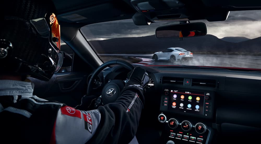 A professional driver is shown behind the wheel of a 2022 Toyota GR 86 driving on a track behind a white car.