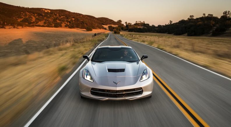 A silver 2014 Chevy Corvette is shown from the front driving on a road after visiting a certified pre-owned Chevy dealer.