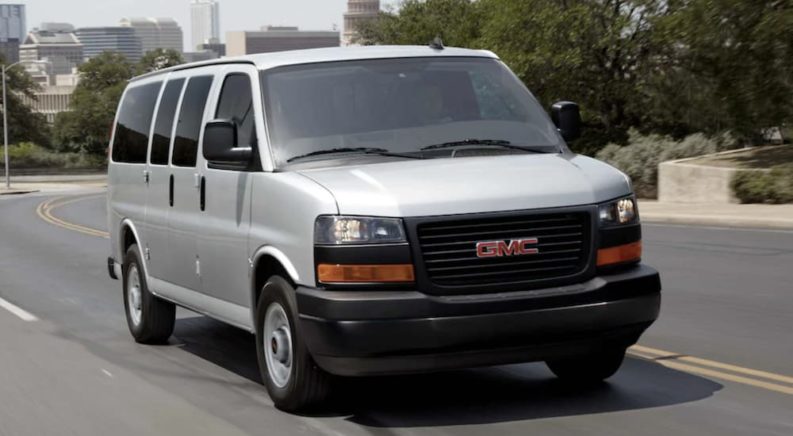 A white 2021 GMC Savana is shown from the front driving on an open road after leaving a used GMC dealership.