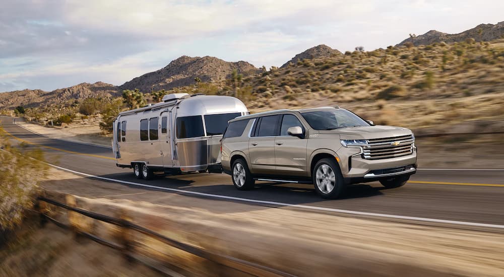 A beige 2022 Chevy Suburban High Country is shown towing an Airstream trailer on an empty highway.