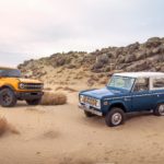A yellow 2021 Ford Bronco and blue 1st Gen Bronco are shown parked in a desert after leaving a Certified Pre-Owned Ford dealership.