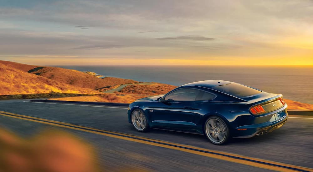 A blue 2021 Ford Mustang is shown from a rear angle driving on a highway with an ocean view.
