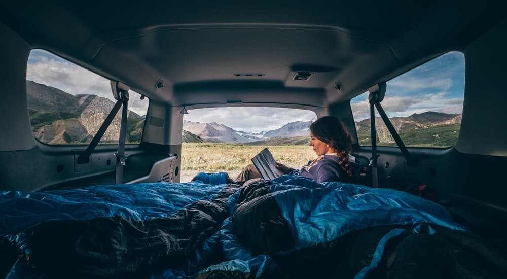 A woman is shown reading a book inside of a van repurposed as a sleeping area.