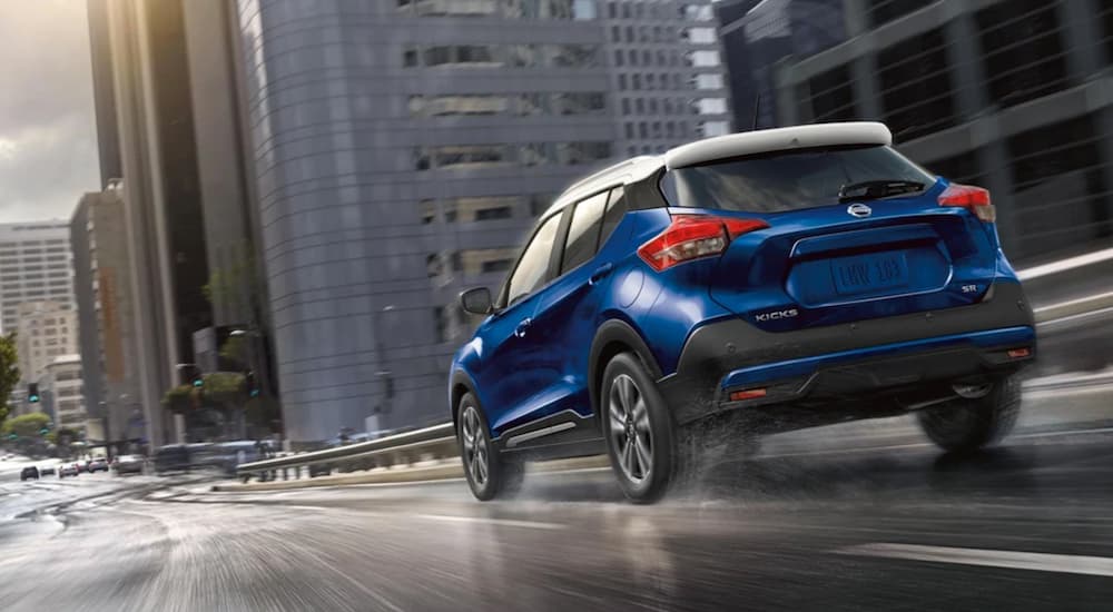 A blue 2020 Nissan Kicks is shown from the rear driving through a city for certified pre-owned Nissan sales.