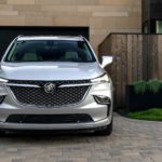 A silver 2022 Buick Enclave is shown from the front parked in front of a modern house during a 2022 Buick Enclave vs 2022 Honda Pilot comparison.