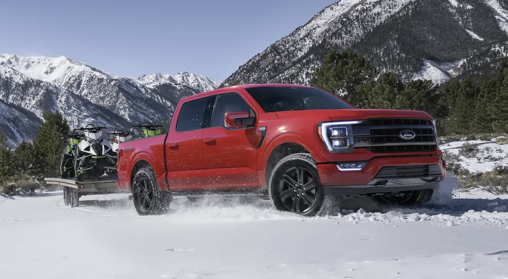 A red 2022 Ford F-150 Limited is shown from the side driving through a snow covered field while pulling a trailer with snowmobiles.