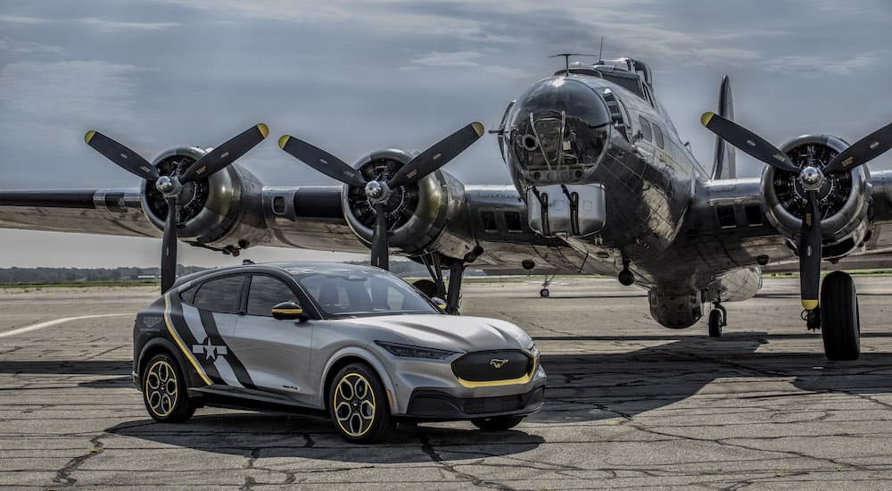 A 2022 Ford Mustang Mach-E is shown from the side in front of a WWII warplane.