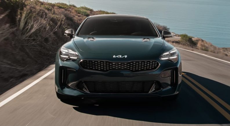 A green 2022 Kia Stinger GT is shown from the front driving on a coastal road after leaving a Kia dealership.