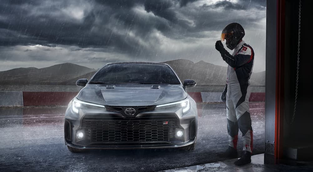 A silver 2023 Toyota GR Corolla is shown from the front in the rain next to a race car driver.