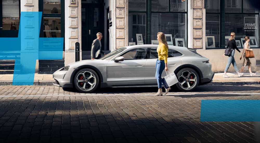 A white 2022 Porsche Tycan is shown parked on a city street next to a person with a shopping bag.